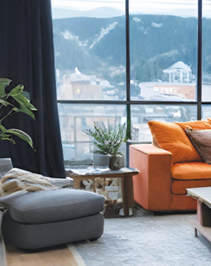 A lounge with a grey ottoman and orange sofa beside floor-to-ceiling windows looking onto hills